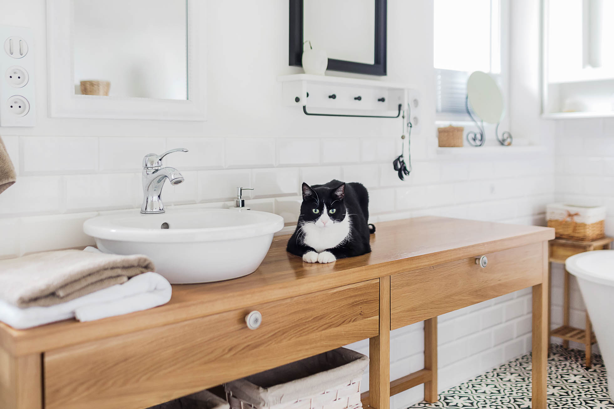 Czarno-biały kot wyleguje się na drewnianym blacie łazienkowym obok białej umywalki. Łazienka jest jasna i czysta, z białymi kafelkami na ścianach i wzorzystą czarno-białą podłogą. Ręczniki są starannie złożone na blacie, a nad umywalką wisi lustro — idealne dla professional fotografia mieszkań i biur in Trójmiasto.