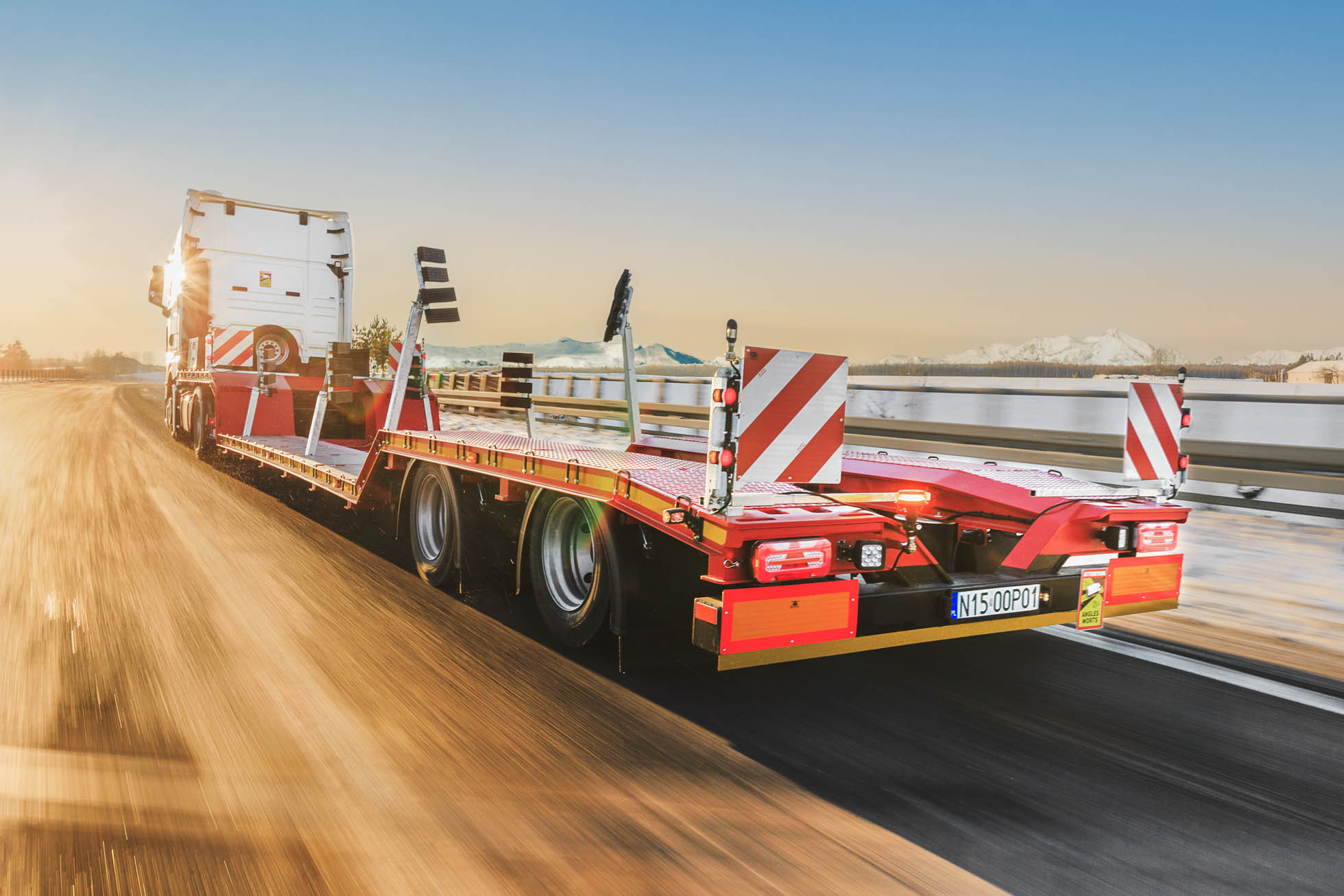 Przyczepa niskopodwoziowa transportowana autostradą o zachodzie słońca, w tle ośnieżone góry. Droga jest pusta, a przyczepa oznaczona czerwonymi i białymi odblaskowymi pasami z dwoma tylnymi barierkami ochronnymi. To zdjęcie uosabia Fotografię komercyjną i motoryzacyjna Trójmiasto, podkreślając jej spokojną, ale potężną istotę.