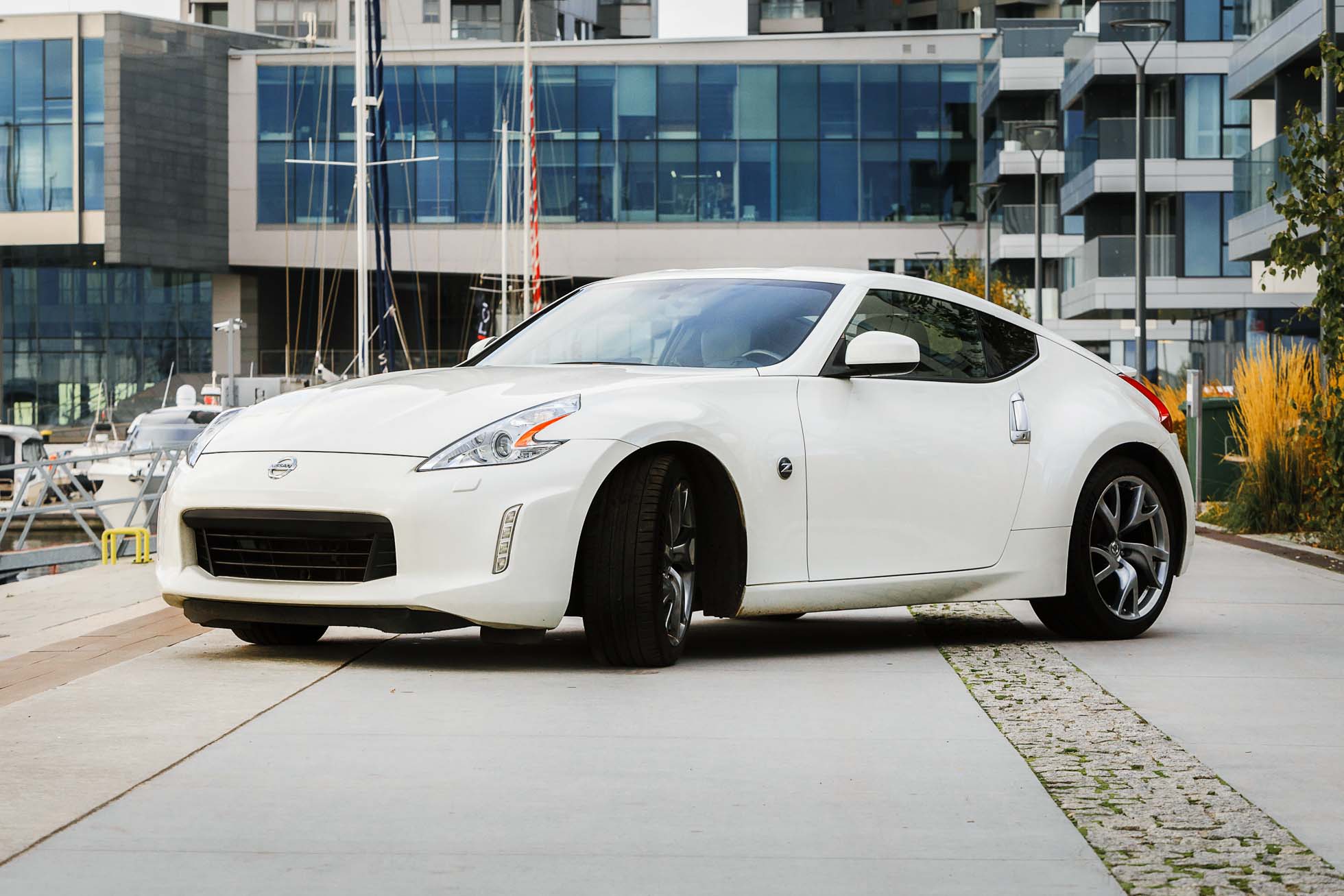 Elegancki biały samochód sportowy zaparkowany na utwardzonej ścieżce w pobliżu mariny, z kilkoma jachtami i żaglówkami w tle. Nowoczesne budynki ze szklanymi fasadami ciągną się wzdłuż nabrzeża, wzmacniając współczesny miejski klimat tego malowniczego widoku Trójmiasta, idealnego do fotografii reklamowej.