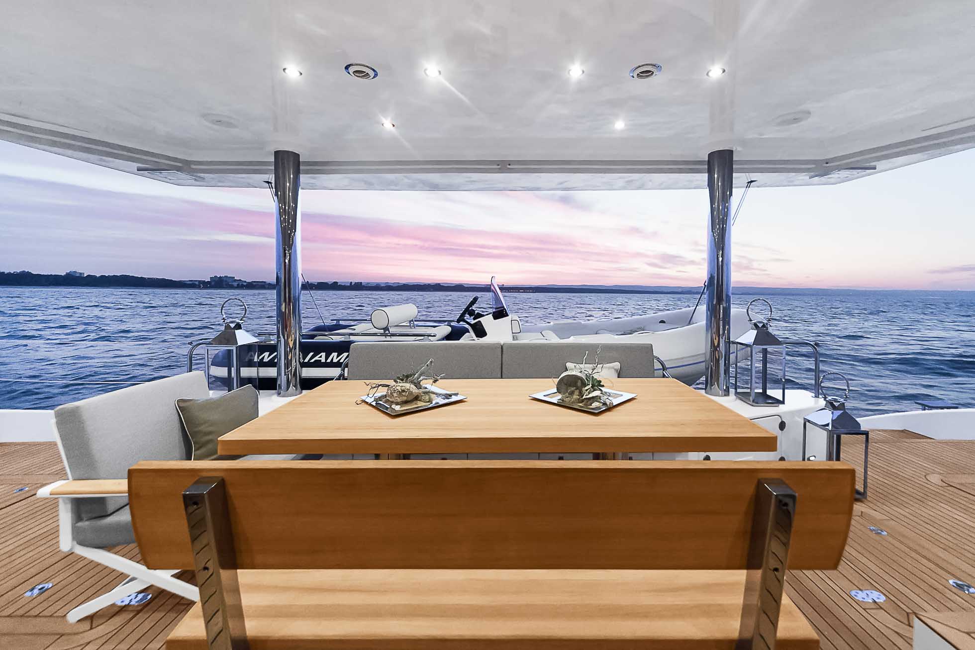 Jadalnia na pokładzie jachtu przeznaczona do spożywania posiłków, z drewnianym stołem i wyściełanymi siedziskami. Wystrój marynistyczny obejmuje latarnie i rozgwiazdy. Jacht znajduje się na spokojnych wodach z malowniczym zachodem słońca w tle, uchwytując istotę profesjonalnej fotografii wnętrz Trójmiasto – najwyższa jakość zdjęć.