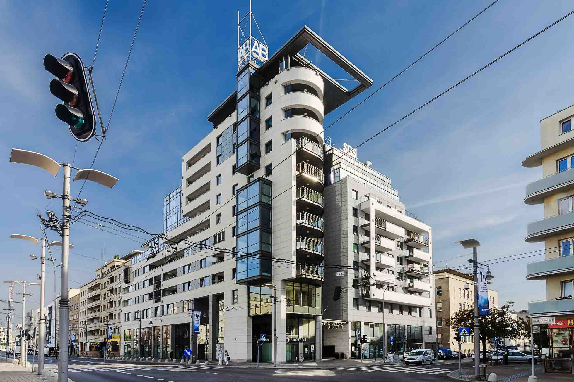 Nowoczesny, wielopiętrowy budynek z mieszanką szkła i białych elementów konstrukcyjnych stoi na skrzyżowaniu. Widoczne są sygnalizacja świetlna i linie energetyczne, a także inne budynki w tle pod czystym, błękitnym niebem. Fotografia wnętrz Trójmiasto – zdjęcia apartamentów i lokali komercyjnych oddają istotę tej miejskiej sceny.