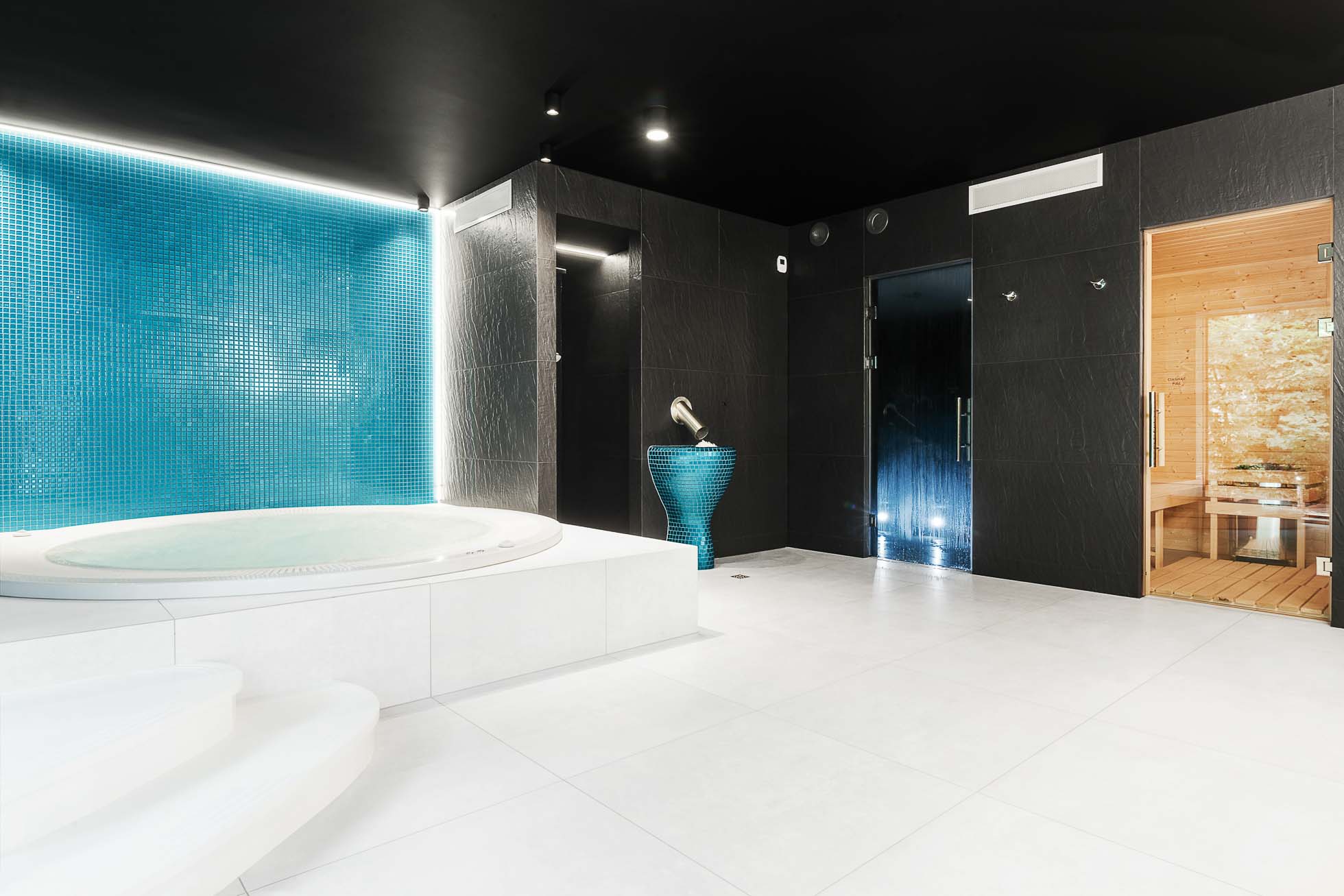 Luksusowy pokój spa z białą okrągłą wanną z hydromasażem na podwyższonej platformie, otoczoną białymi kafelkami. Obok znajduje się żywa turkusowa ściana mozaikowa, z nowoczesną czarną drewnianą sauną wyposażoną w szklane drzwi po prawej stronie. Przestrzeń ma elegancki, współczesny wystrój. Idealny dla fotografia wnętrz Trójmiasto – zdjęcia spa i hoteli.