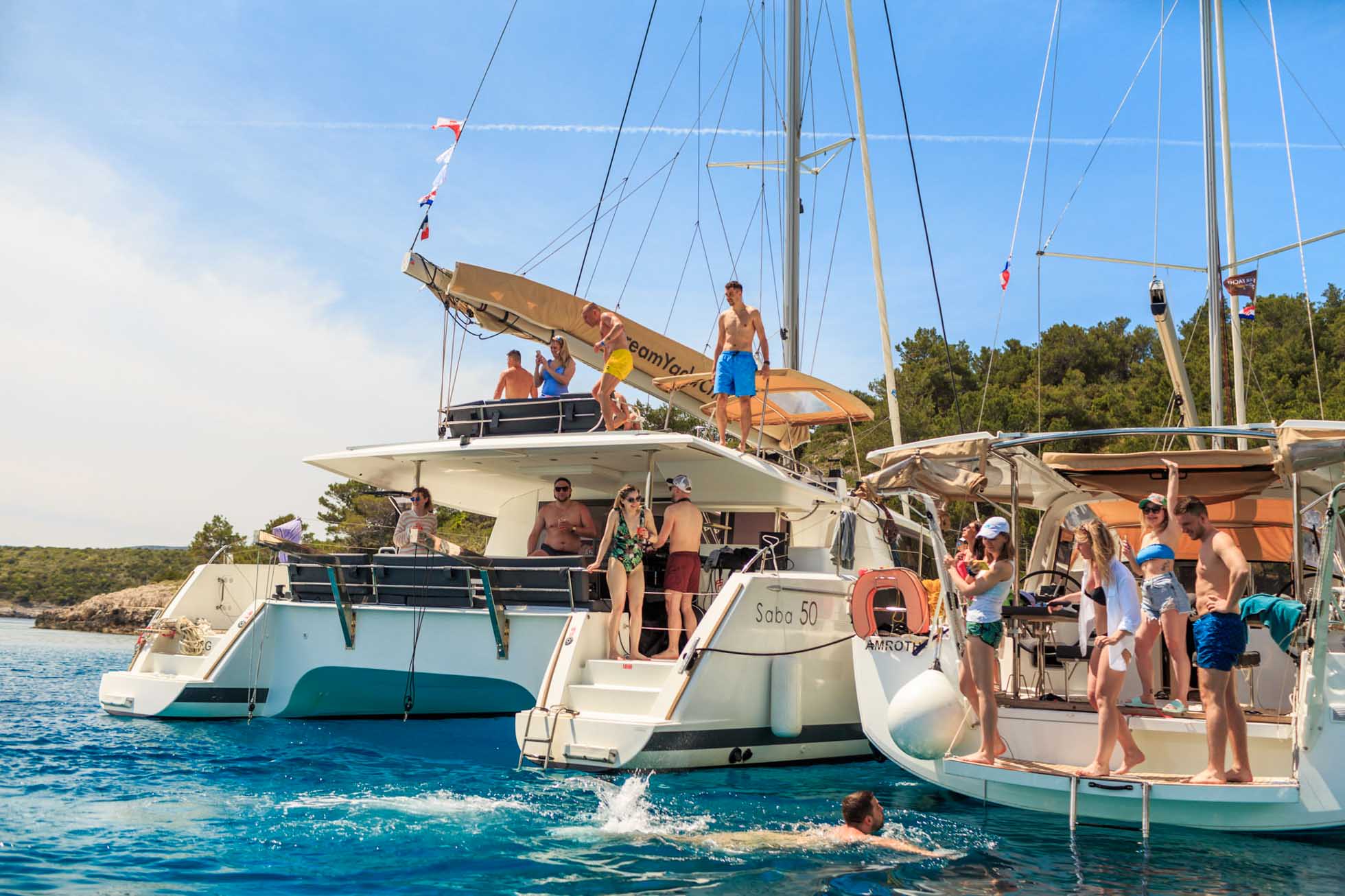 Grupa ludzi ciesząca się słonecznym dniem na dwóch zakotwiczonych żaglówkach na czystym, błękitnym morzu. Niektórzy stoją na pokładach, podczas gdy inni pływają lub relaksują się w wodzie, uwieczniając chwile idealne dla Fotografia lifestyle Trójmiasto. W tle zielona, zalesiona linia brzegowa pod jasnoniebieskim niebem.