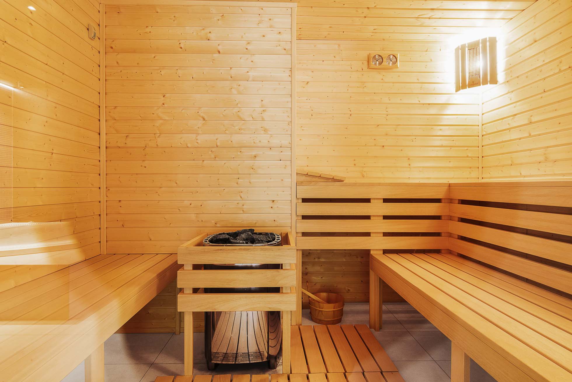 Nowoczesna drewniana sauna z panelowymi ścianami i ławkami. W rogu znajduje się piec z kamieniami na górze, a na podłodze drewniane wiadro i chochla. Ciepłe, miękkie oświetlenie rozświetla przestronne wnętrze, tworząc relaksującą atmosferę. Profesjonalna fotografia wnętrz Trójmiasto pięknie uchwyciła tę spokojną przestrzeń.