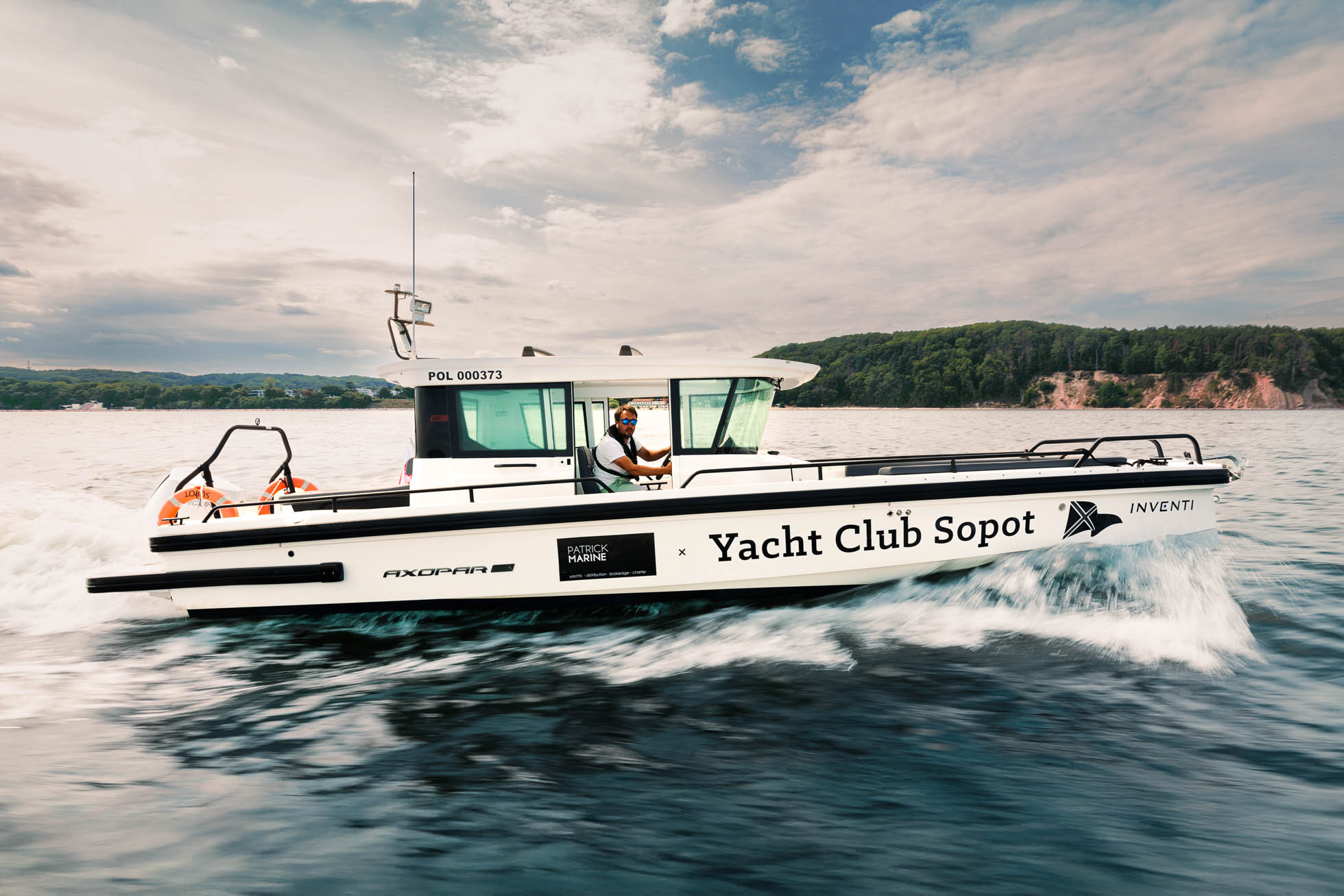 Biały jacht z napisem „Yacht Club Sopot” na boku żegluje po zatoce gdanskiej, idealnym na sesje wizerunkowe dla stoczni jachtowych Gdańsk. Za sterami widać osobę. W tle bujne zielone wzgórza pod częściowo zachmurzonym niebem. Jacht porusza się szybko, tworząc fale za sobą.