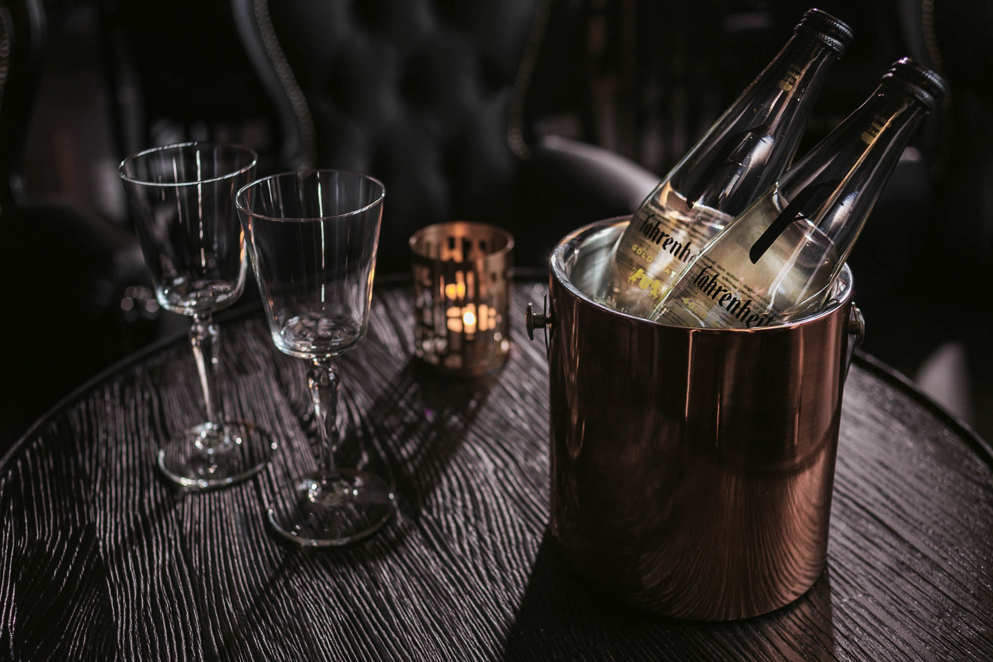 scena z dwiema butelkami wody chłodzącymi się w metalowym wiaderku z lodem ustawionym na ciemnym drewnianym stole. W pobliżu znajdują się dwa puste kieliszki i mały, świecący świecznik, tworzący przytulną atmosferę. W tle znajdują się pluszowe, ciemne krzesła — idealne dla Fotografia reklamowa Trójmiasto.