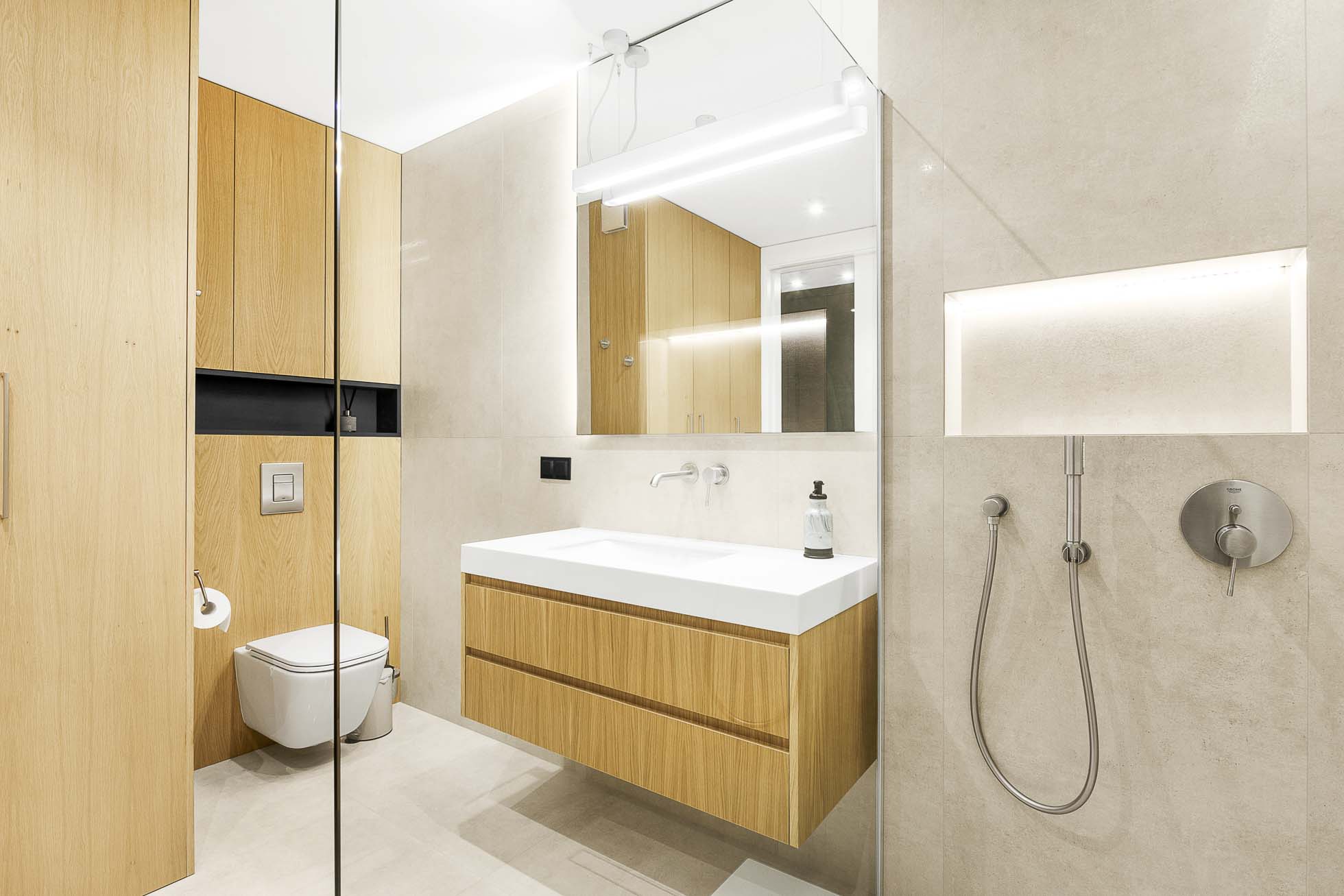 Nowoczesna łazienka z jasnymi drewnianymi szafkami, wiszącą toaletą i wiszącą toaletką z białym blatem i umywalką. Nad umywalką znajduje się duże lustro z oprawą oświetleniową. Prysznic ma deszczownicę i wnękę, doskonale uchwyconą w Fotografia wnętrz Trójmiasto – zdjęcia apartamentów i lokali komercyjnych. Ogólny projekt jest minimalistyczny.