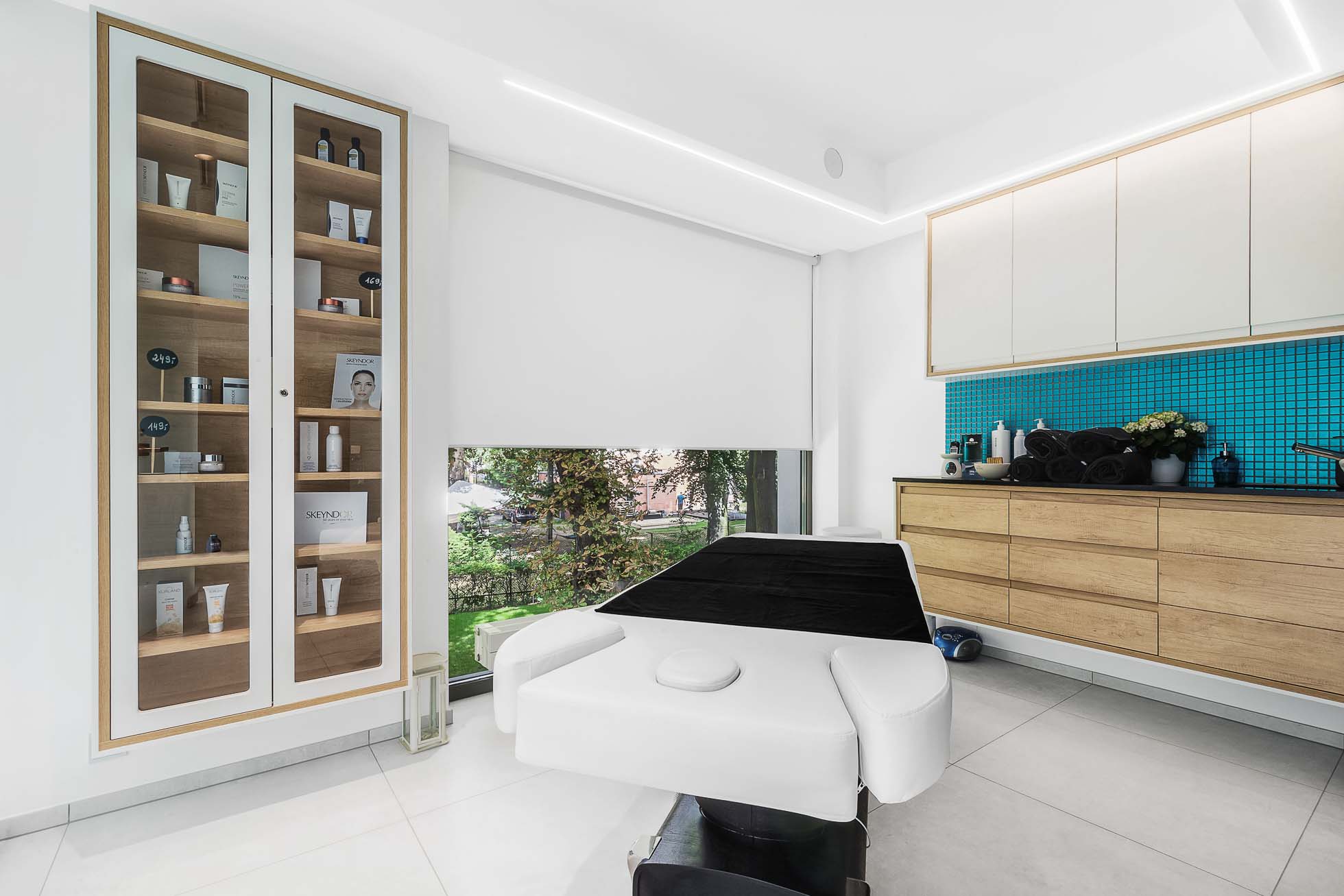 Nowoczesny gabinet zabiegowy lub spa z białym stołem zabiegowym, drewnianymi szafkami i półkami eksponującymi różne produkty. Duże okno z widokiem na ogród jest częściowo zasłonięte białą żaluzją. Obszar zabiegowy zdobią niebieskie mozaiki, ukazujące istotę Sopotu – usługi fotograficzne.