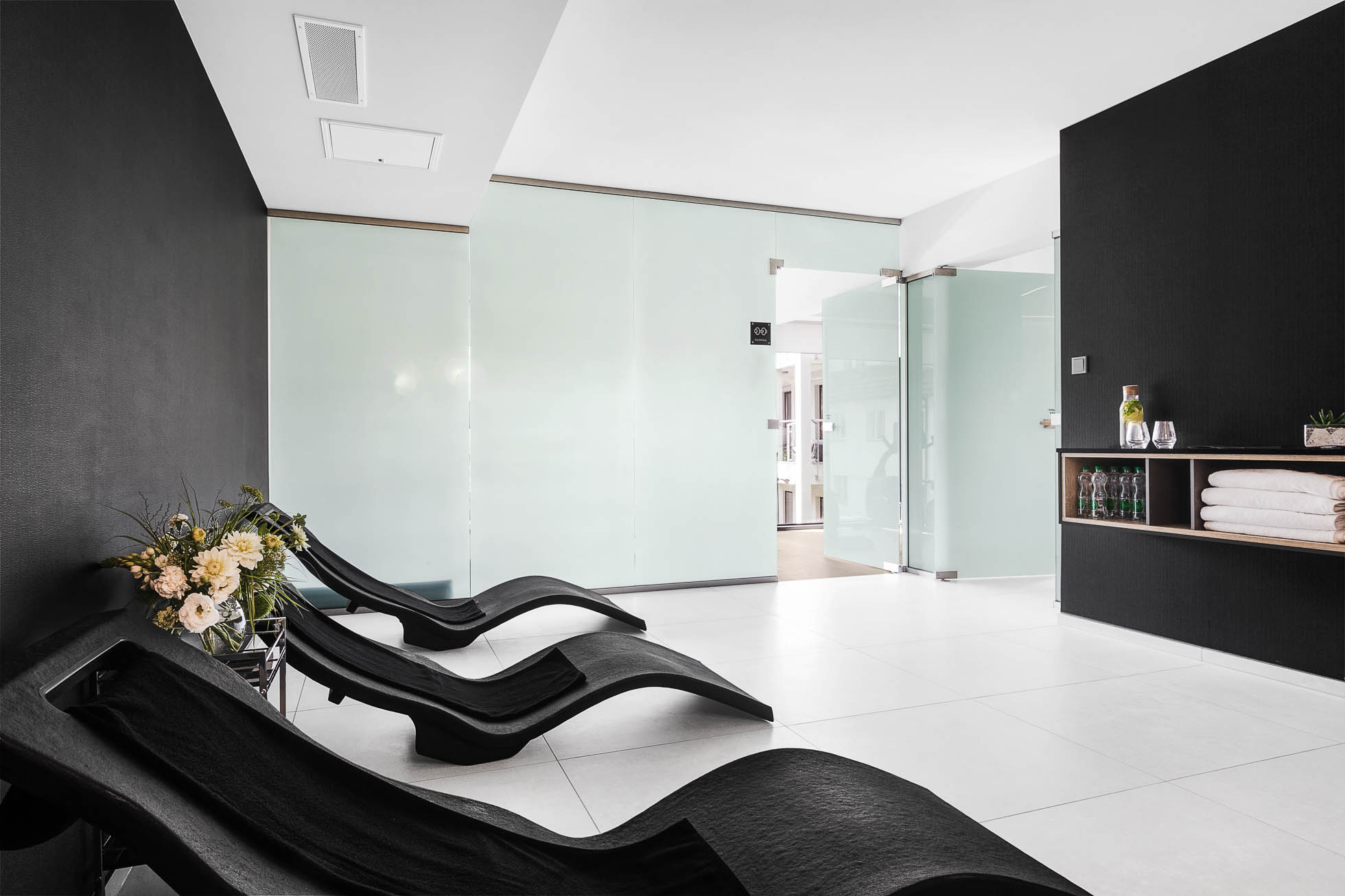 Nowoczesny pokój relaksacyjny spa w Trójmieście z trzema czarnymi ergonomicznymi fotelami wypoczynkowymi. Minimalistyczny design obejmuje jasną podłogę i ściany z matowego szkła. Mały stolik mieści ręczniki, wodę i kwiaty, dodając przestrzeni komfortu i świeżości, idealny dla Fotografia architektury Trójmiasto.