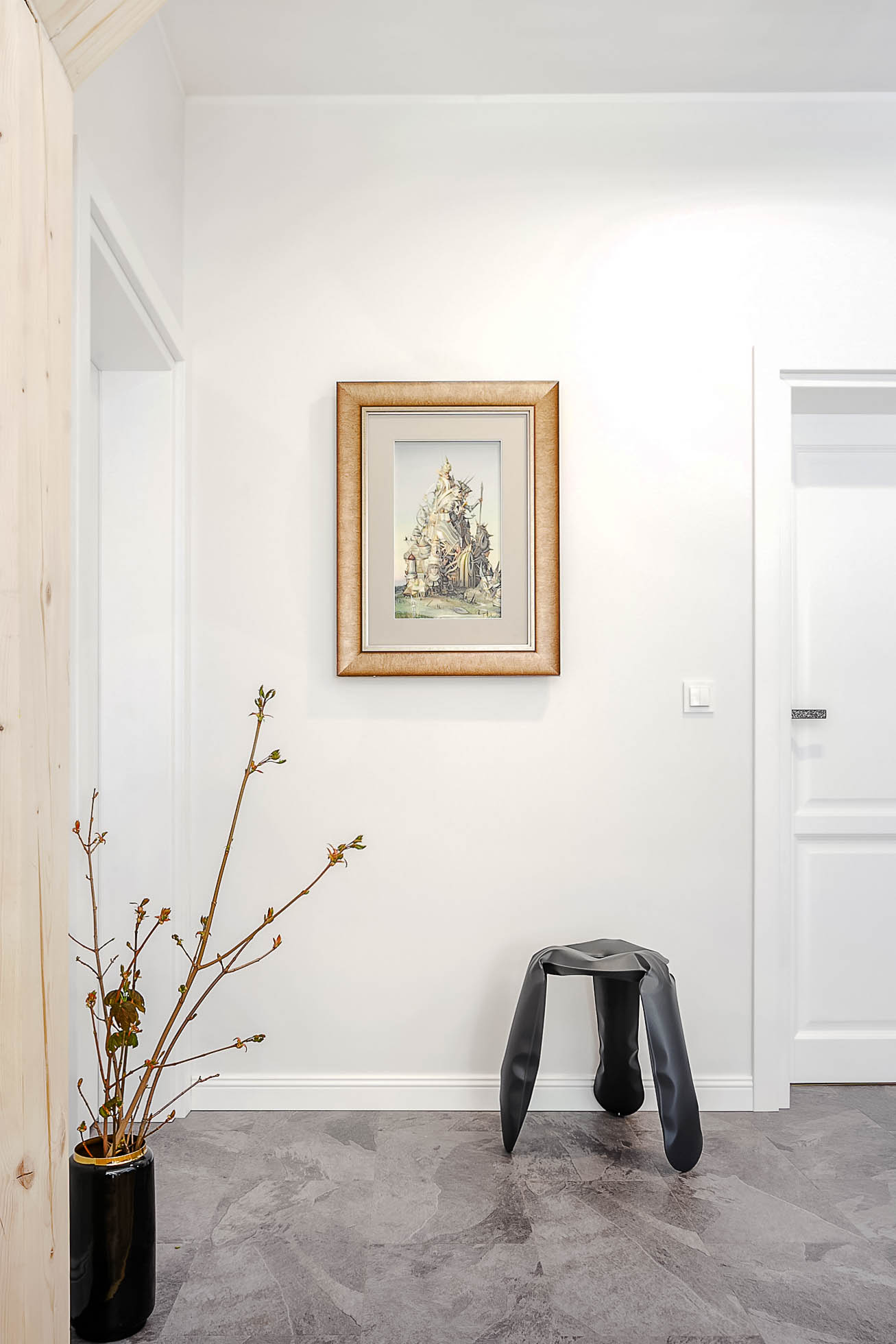Minimalistyczny korytarz ma jasnoszarą podłogę, białe ściany i białe drzwi. Na ścianie wisi oprawiony obraz. Po prawej stronie stoi czarny artystyczny stołek, a po lewej czarna doniczka z gałęziami. Fotografia wnętrz Trójmiasto – zdjęcia apartamentów i lokali komercyjnych bezproblemowo uchwyca takie eleganckie przestrzenie.