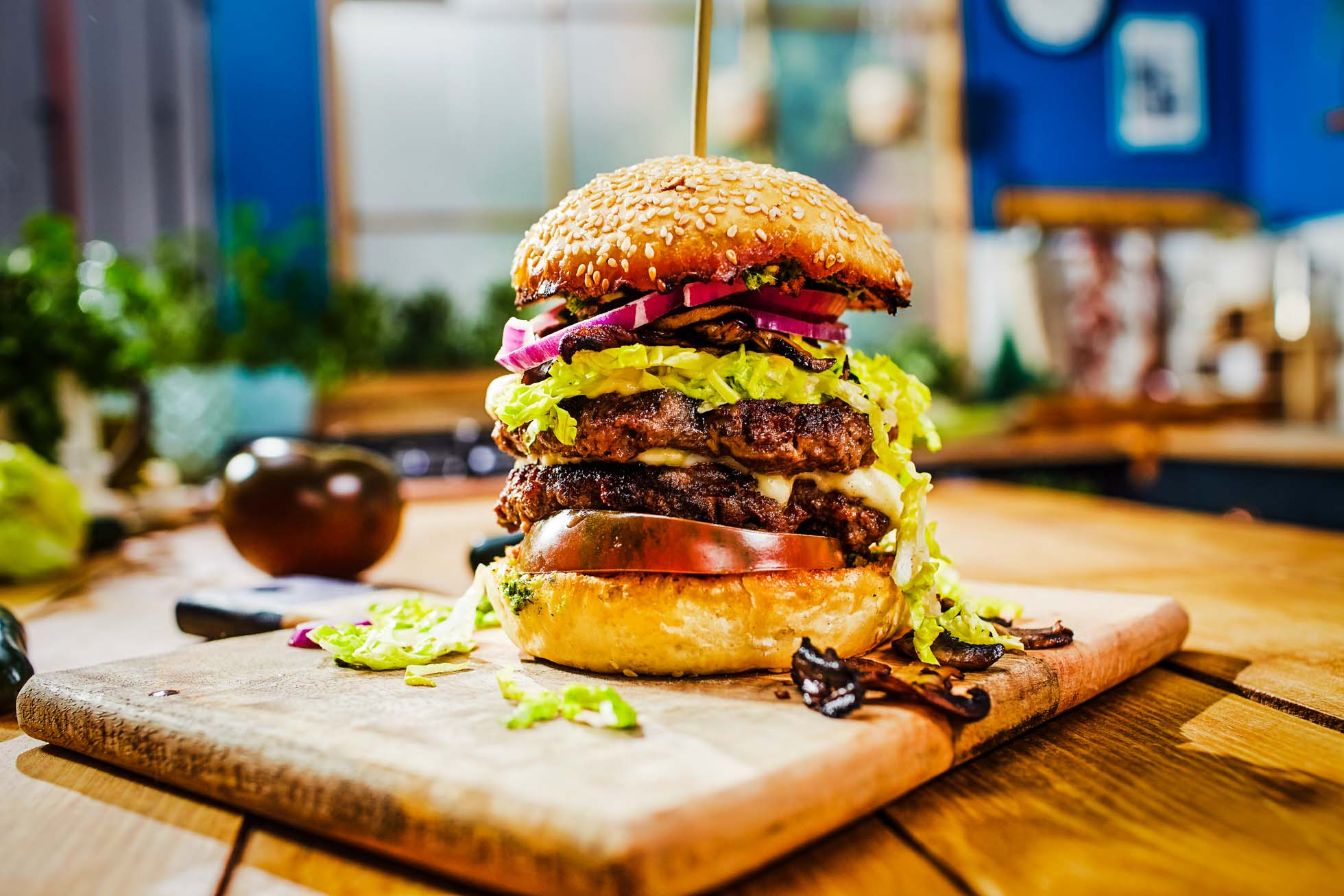 Soczysty podwójny cheeseburger z sałatą, pomidorem, roztopionym serem, ogórkami kiszonymi i cebulą w bułce z sezamem jest serwowany na drewnianej desce do krojenia. Burger jest ozdobiony szpikulcem i doskonale uchwycony jako część Fotografia reklamowa Trójmiasto w jasno oświetlonej kuchni z kolorowym wystrojem.