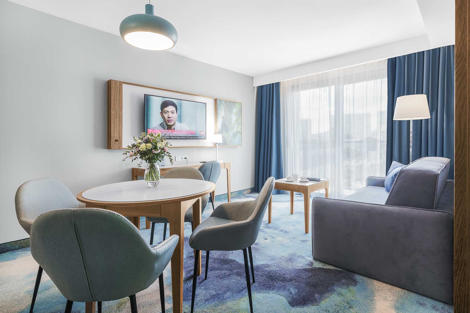 Nowoczesny salon w Trójmieście wyposażony jest w okrągły stół jadalny z czterema krzesłami, kanapę, telewizor zamontowany na ścianie i duże okno z niebieskimi zasłonami. Udekorowany w odcieniach niebieskiego i szarości, zawiera wiszącą lampę i kwiatowy element centralny. Idealny do fotografia architektury Trójmiasto – zdjęcia wnętrz i hoteli.