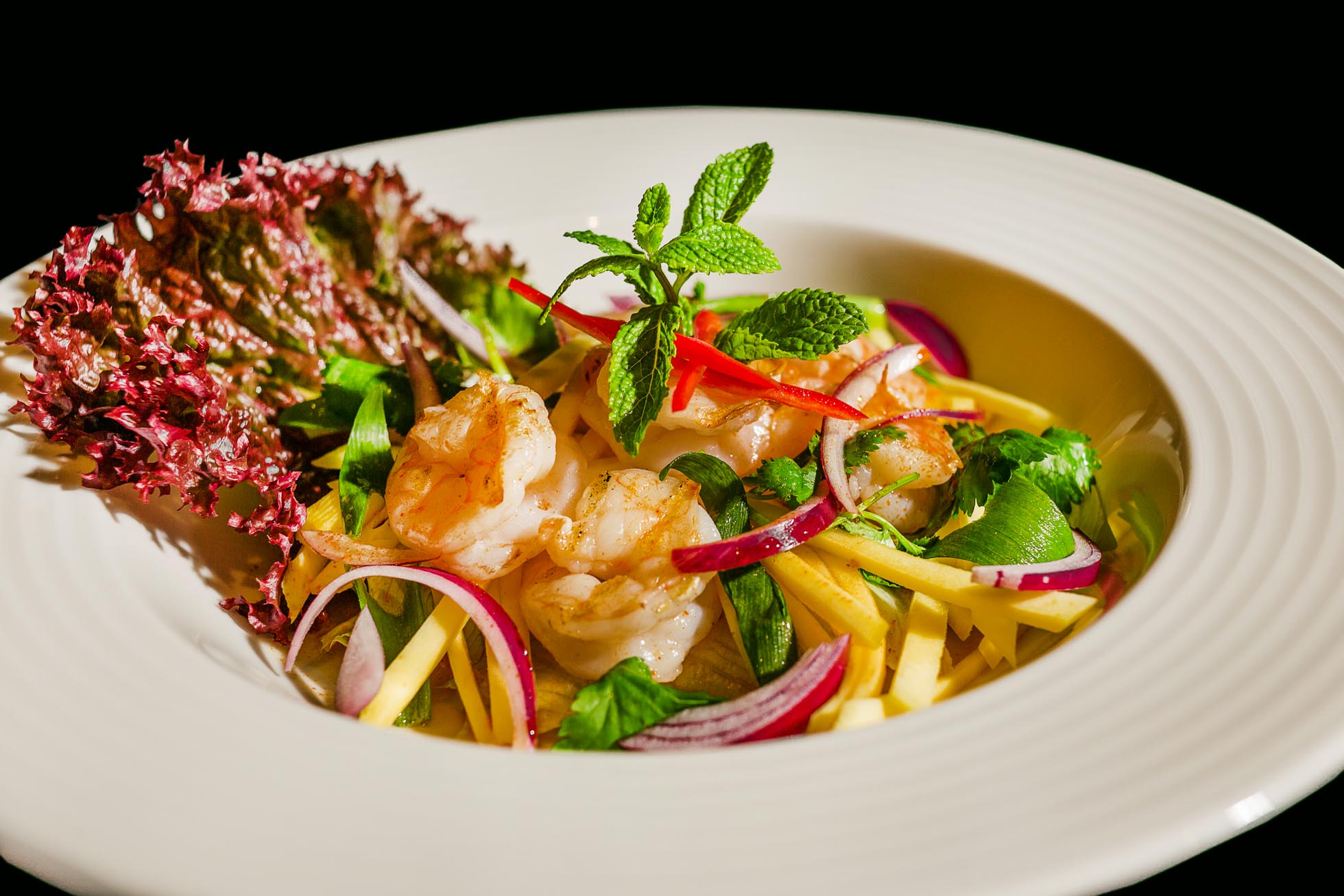 Żywa sałatka z krewetkami, plasterkami czerwonej cebuli, paskami żółtej papryki, zielonymi liśćmi i listkami mięty, ozdobiona gałązką mięty i czerwoną sałatą. To oszałamiające danie jest prezentowane w białej misce na czarnym tle — idealne dla Fotografia dla restauracji Trójmiasto.