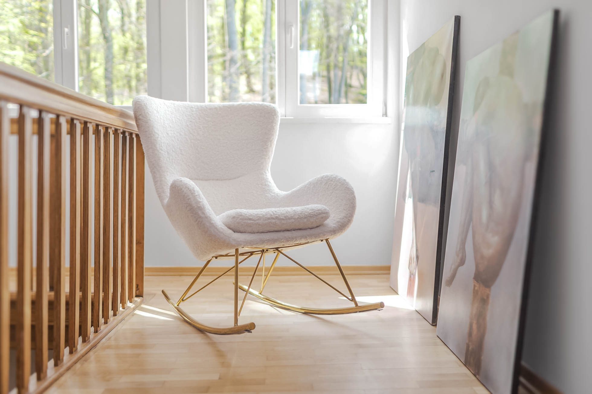 Przytulny biały fotel bujany ze złotą ramą stoi przy jasnym oknie w minimalistycznym, słonecznym pokoju z jasną drewnianą podłogą. Dwa duże, pochylone płótna sztuki abstrakcyjnej są umieszczone na podłodze przy ścianie obok fotela, idealne do Fotografia wnętrz Trójmiasto – zdjęcia apartamentów i lokali komercyjnych.