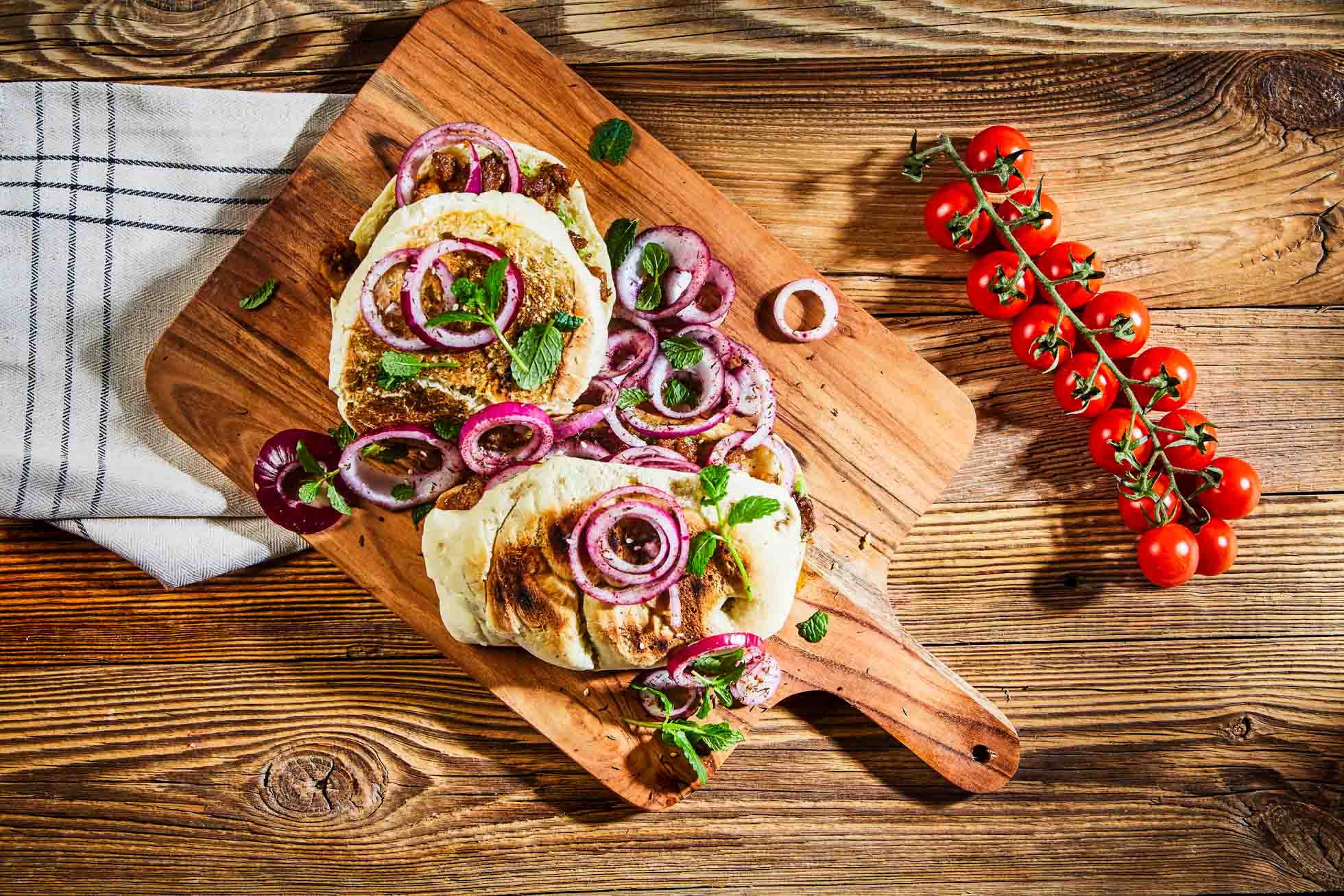 Drewniana deska do krojenia z plastrami grillowanego sera halloumi, zwieńczona krążkami czerwonej cebuli i ziołami. Obok deski znajduje się kratkowana ściereczka kuchenna i pęczek dojrzałych na krzaku pomidorków koktajlowych na rustykalnym drewnianym stole — idealne do fotografia produktowa i kulinarna Gdańsk.