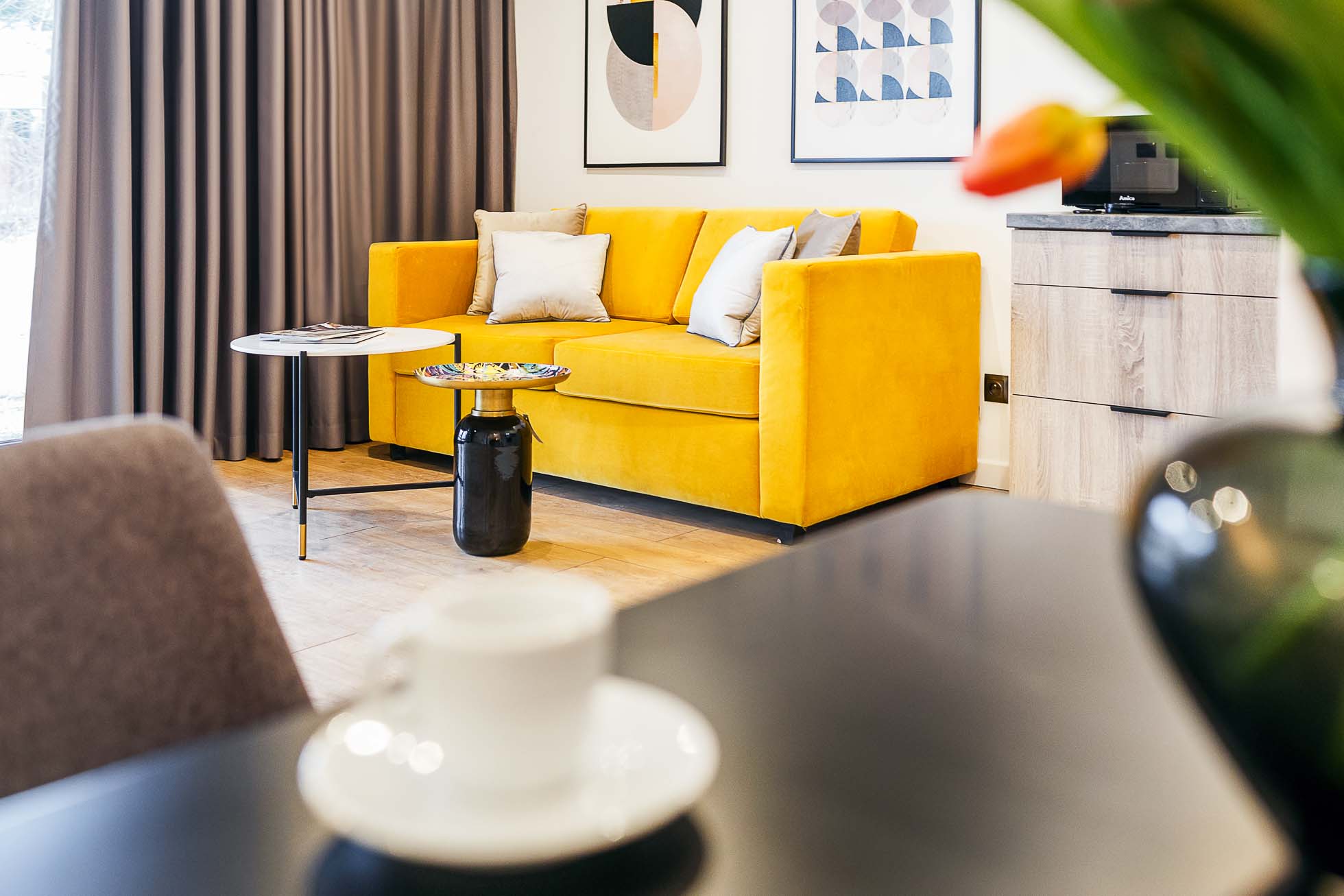 Przytulny salon w Gdyni z żywą żółtą sofą ozdobioną poduszkami, małym czarnym stolikiem bocznym z miską, minimalistyczną sztuką ścienną i brązową zasłoną. Na pierwszym planie znajduje się stolik z białą filiżanką i spodkiem oraz rozmyta zielona roślina z pomarańczowym kwiatem — idealna dla miłośników Fotografia architektury Trójmiasto.