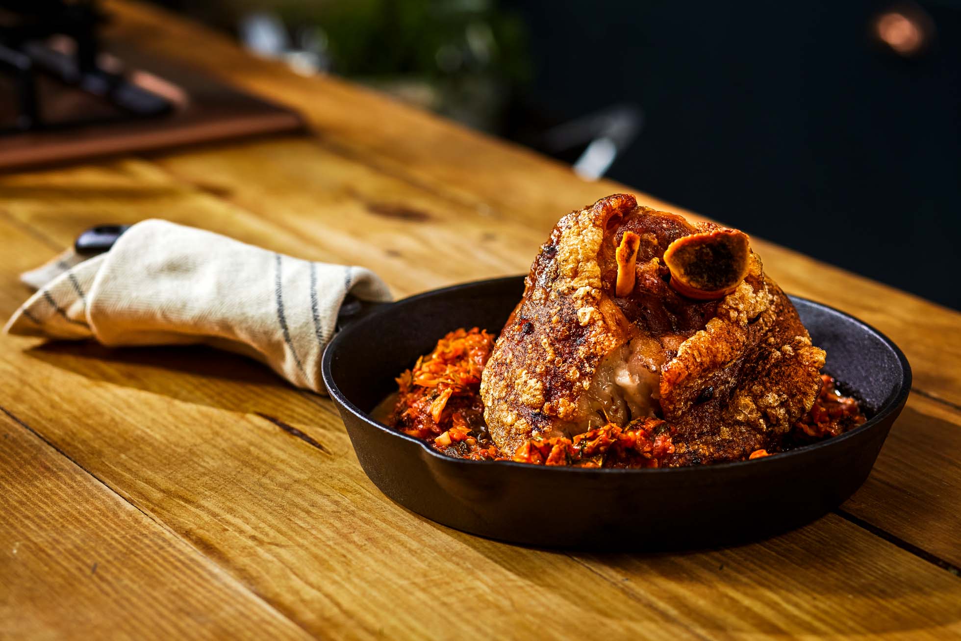 Smaczna pieczona golonka wieprzowa leży na czarnej patelni na drewnianym stole. Zrumieniona, chrupiąca skórka lśni, a całość leży na drobno posiekanych warzywach. Na rączce patelni leży biały ręcznik kuchenny z ciemnym paskiem, idealny do fotografia kulinarna Trójmiasto.