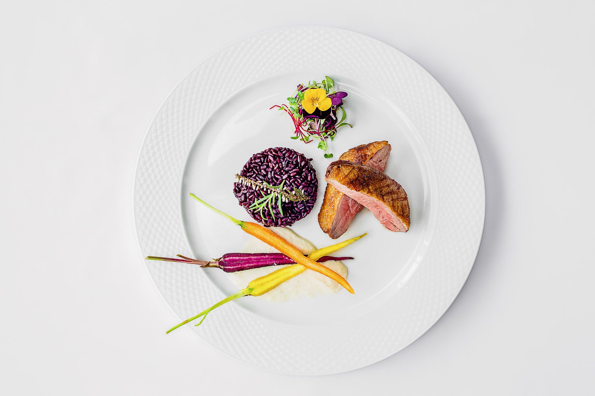 Biały talerz z elegancko ułożonym posiłkiem z pokrojoną piersią kaczki, czarnym ryżem, sałatką z jadalnymi kwiatami i marchewkami heirloom. Prezentacja jest artystyczna i wyrafinowana na jednolitym białym tle — idealna dla miłośników fotografii produktowej i kulinarnej Gdańsk.