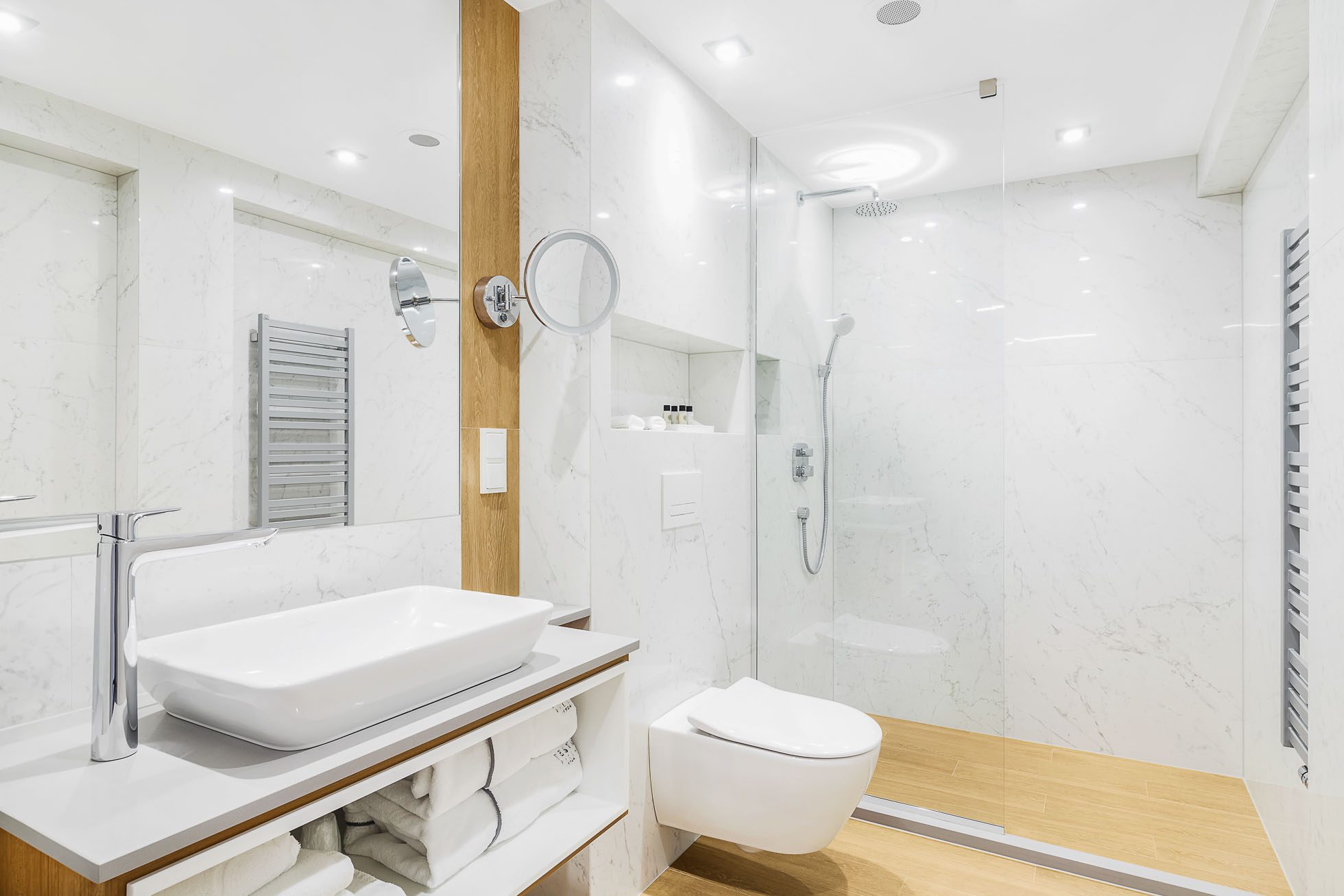 Nowoczesna, jasna łazienka z białymi marmurowymi ścianami i jasną drewnianą podłogą. Posiada elegancką prostokątną umywalkę nablatową, wiszącą toaletę, dużą kabinę prysznicową ze szklanymi drzwiami, półkę na ręczniki z dokładnie złożonymi ręcznikami i oświetlenie wnękowe. Idealna dla Fotografia wnętrz Trójmiasto – zdjęcia apartamentów i lokali komercyjnych w Gdyni.