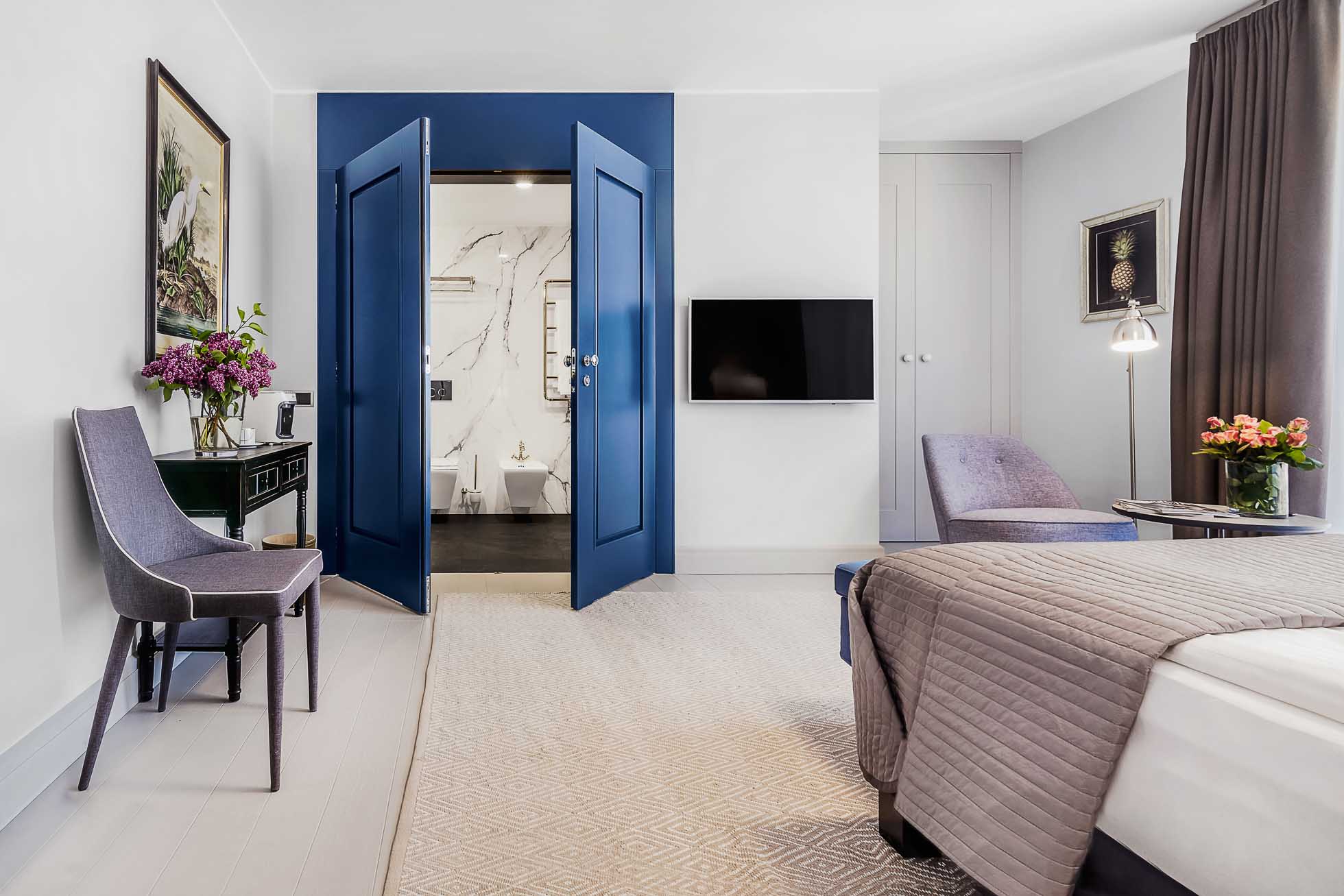 Nowoczesny pokój hotelowy o przestronnym układzie, wyposażony w łóżko z szarą kołdrą, biurko i krzesło oraz leżankę z stolikiem nocnym. W pokoju znajduje się telewizor zamontowany na ścianie i podwójne niebieskie drzwi prowadzące do łazienki z akcentami marmurowymi. Wystrój obejmuje kompozycje kwiatowe i oprawione dzieła sztuki. Fotografia wnętrz Trójmiasto – zdjęcia apartamentów i hoteli
