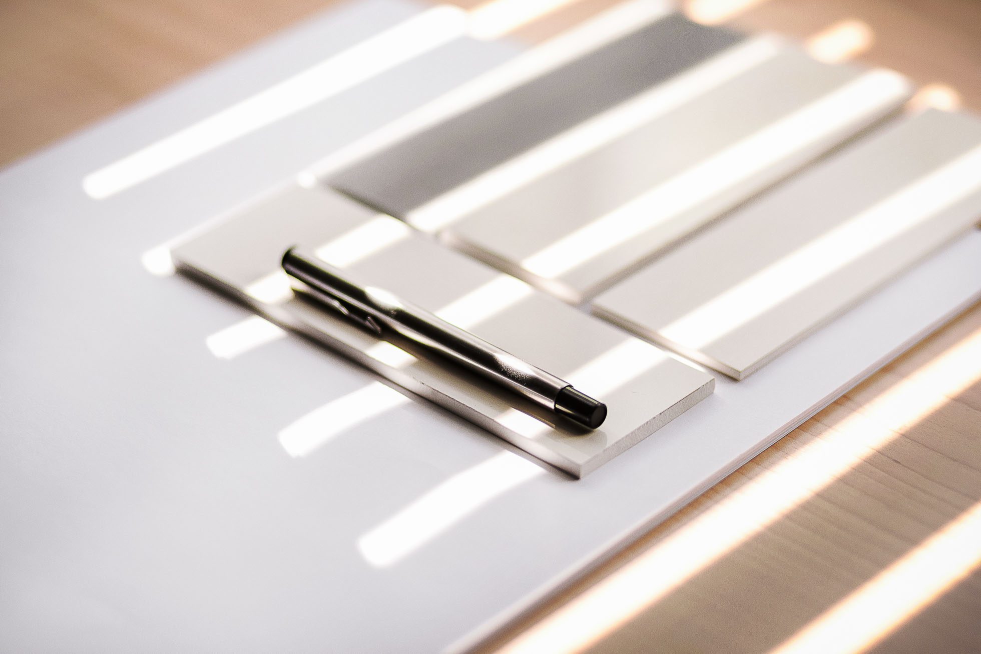 Czarny długopis leży poziomo na czterech prostokątnych próbkach farby o różnych odcieniach bieli i szarości, ułożonych pionowo na białej powierzchni. Tło, delikatnie oświetlone światłem słonecznym wpadającym przez żaluzje i rzucającym prążkowane cienie, emanuje wyrafinowanym akcentem widocznym w sesjach fotograficznych wnętrz Gdańsk.