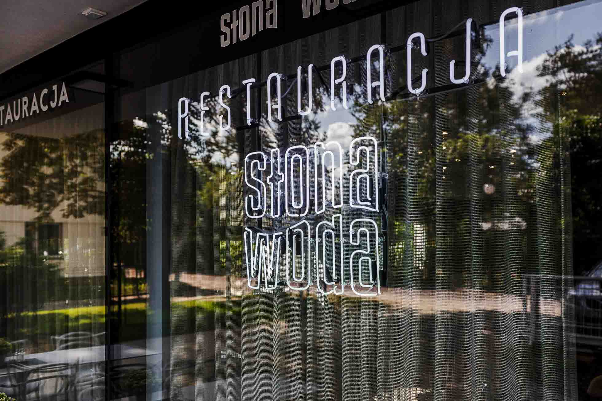 Nowoczesna fasada restauracji z dużymi szklanymi oknami wyświetla podświetlony napis „Restauracja Słona Woda”. Odbicia drzew i miejsc siedzących na zewnątrz można zobaczyć na szkle, tworząc połączenie wnętrza i otoczenia zewnętrznego, doskonale uchwycone przez Fotografia architektury Trójmiasto – restauracji i hoteli.