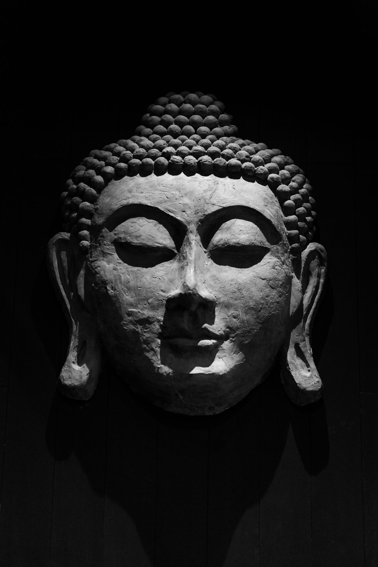 Czarno-białe zdjęcie spokojnej rzeźby głowy Buddy z zamkniętymi oczami i szczegółowymi kręconymi włosami na ciemnym tle. Duże, wydłużone płatki uszu rzeźby przekazują spokój i ciszę, idealne do sesji fotograficznych wnętrz Gdańsk lub uchwycenia spokoju potrzebnego w profesjonalnej fotografii.