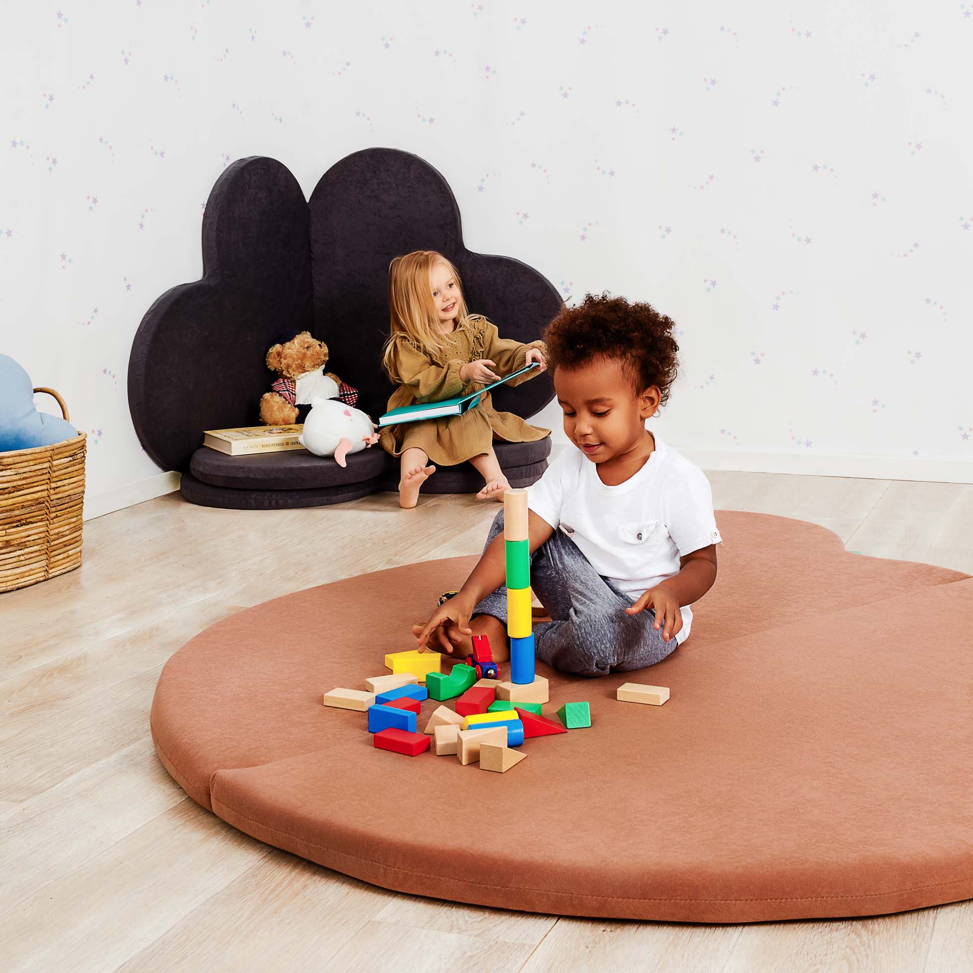 Dwoje małych dzieci jest w pokoju zabaw. Jedno dziecko siedzi na brązowej, wyściełanej macie w kształcie chmury i bawi się kolorowymi drewnianymi klockami. Drugie dziecko siedzi na czarnym, wyściełanym siedzeniu w kształcie chmury i patrzy na książkę. W pobliżu znajduje się kosz z zabawkami i pluszowymi zwierzętami — idealny do fotografia reklamowa Trójmiasto.