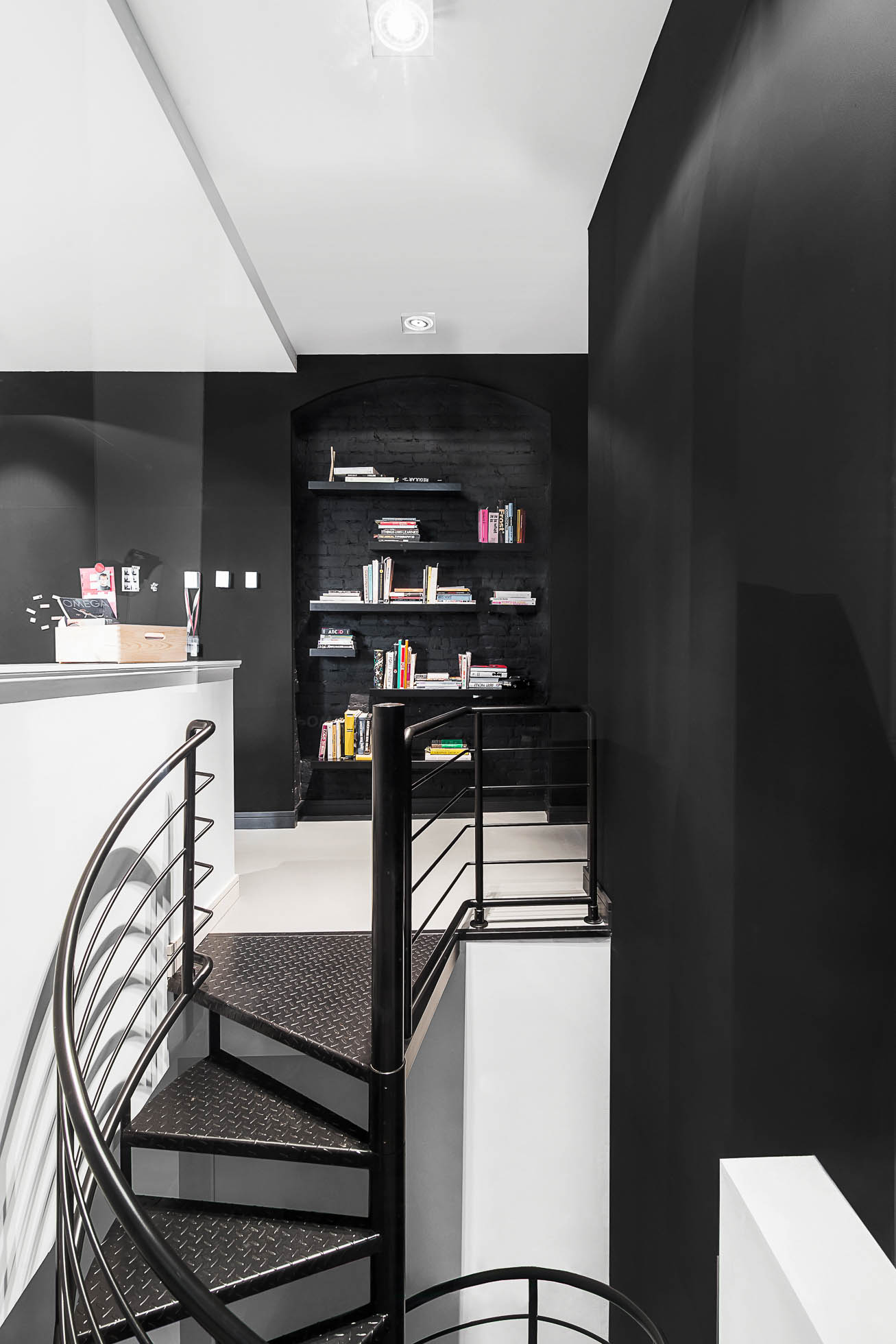 Nowoczesne wnętrze w Gdyni z czarnymi spiralnymi schodami prowadzącymi na wyższy poziom. Ściany pomalowane na czarno i biało. W tle widać wbudowaną półkę na książki osadzoną w czarnej wnęce, wypełnioną różnymi książkami i przedmiotami dekoracyjnymi.