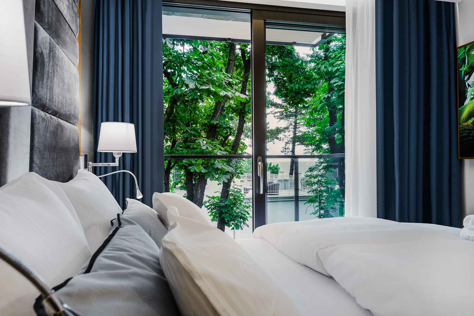 Na zdjęciu nowoczesna sypialnia z dużym łóżkiem, puszystą białą pościelą i wieloma poduszkami. Obok łóżka stoi stolik nocny z zapaloną lampą. Okna od podłogi do sufitu ukazują zielone liście oprawione w ciemnoniebieskie zasłony, pięknie uchwycone przez Fotografia wnętrz Trójmiasto – zdjęcia apartamentów i hoteli