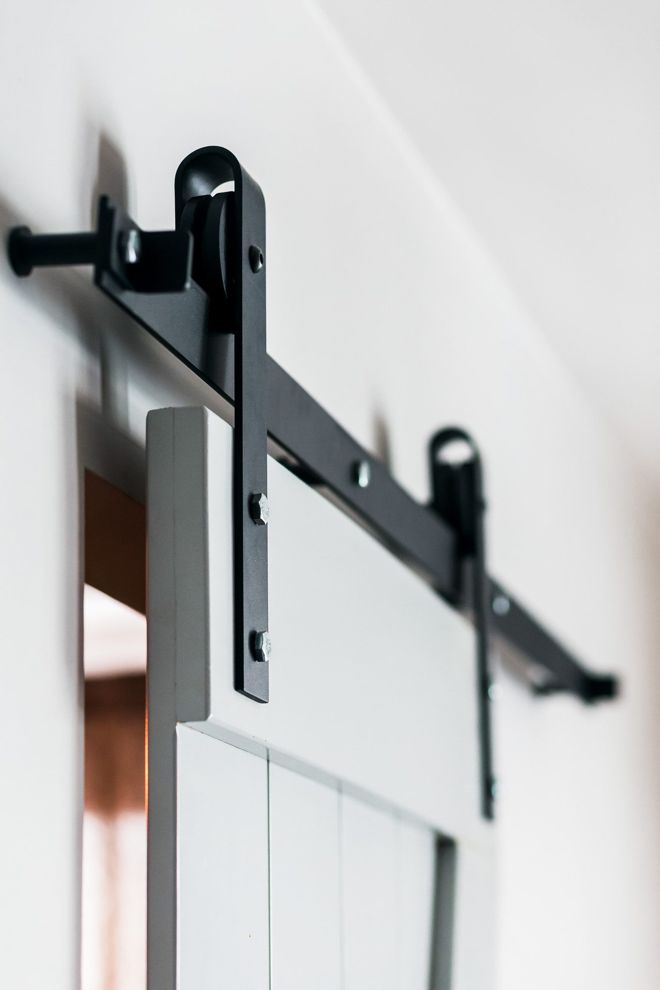 Zbliżenie na przesuwane drzwi do stodoły zamontowane na czarnym metalowym systemie szyn. Drzwi są częściowo otwarte, odsłaniając górny róg i prezentując nowoczesny design przemysłowy okuć na tle białej ściany. Oto 6 fraz SEO Fotografia wnętrz Trójmiasto – zdjęcia apartamentów i lokali komercyjnych.