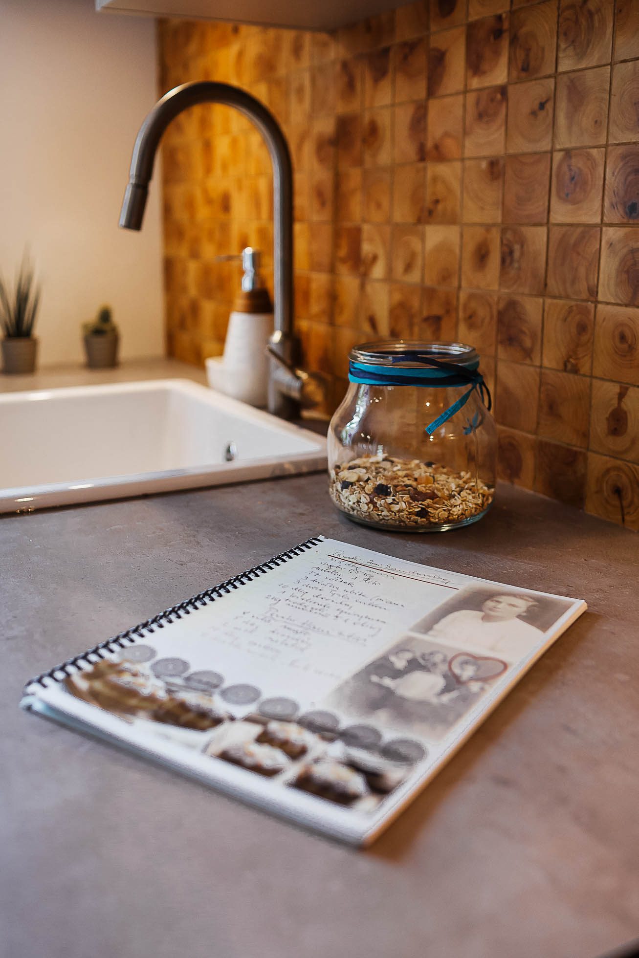 Blat kuchenny ze zlewem, doniczkowymi roślinami i słoikiem wypełnionym ziarnami. Na pierwszym planie otwarty spiralny notatnik wyświetla przepis obok zdjęcia kobiety. Panel przeciwbryzgowy składa się z kwadratowych płytek o wzorze drewna – idealnych do sesje fotograficzne wnętrz Gdańsk,Gdynia,Sopot usługi fotograficzne.