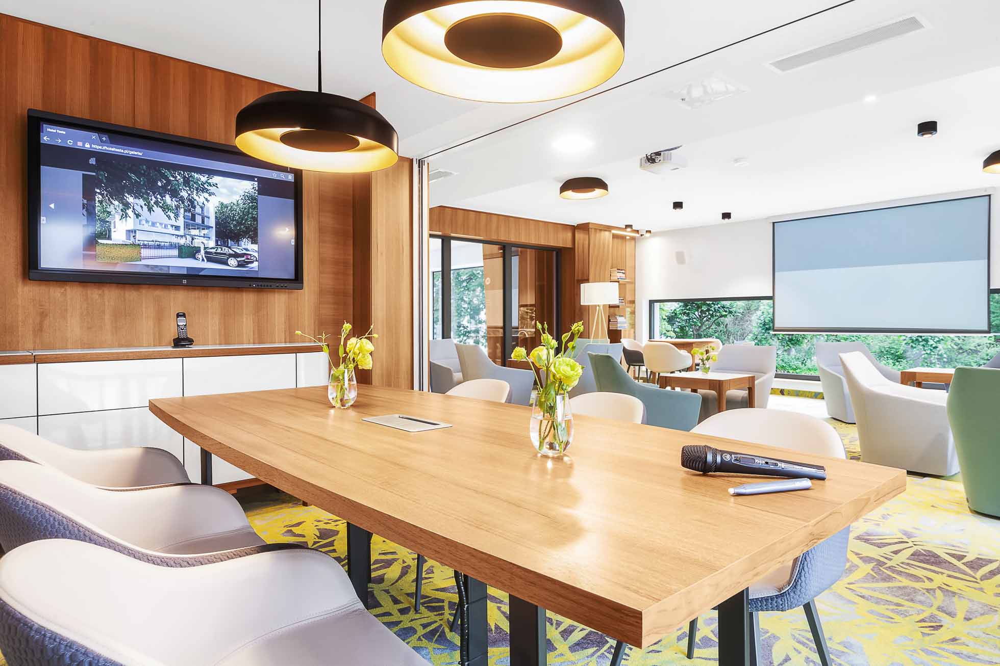 Nowoczesna sala konferencyjna z dużym prostokątnym drewnianym stołem, otoczonym krzesłami. W sali znajduje się zamontowany na ścianie ekran wyświetlający prezentację, oświetlenie sufitowe i duże okno z widokiem na zieleń. Na stole stoją wazony z żółtymi kwiatami. Fotografia architektury Trójmiasto – zdjęcia wnętrz i budynków naprawdę oddaje elegancję tej przestrzeni.