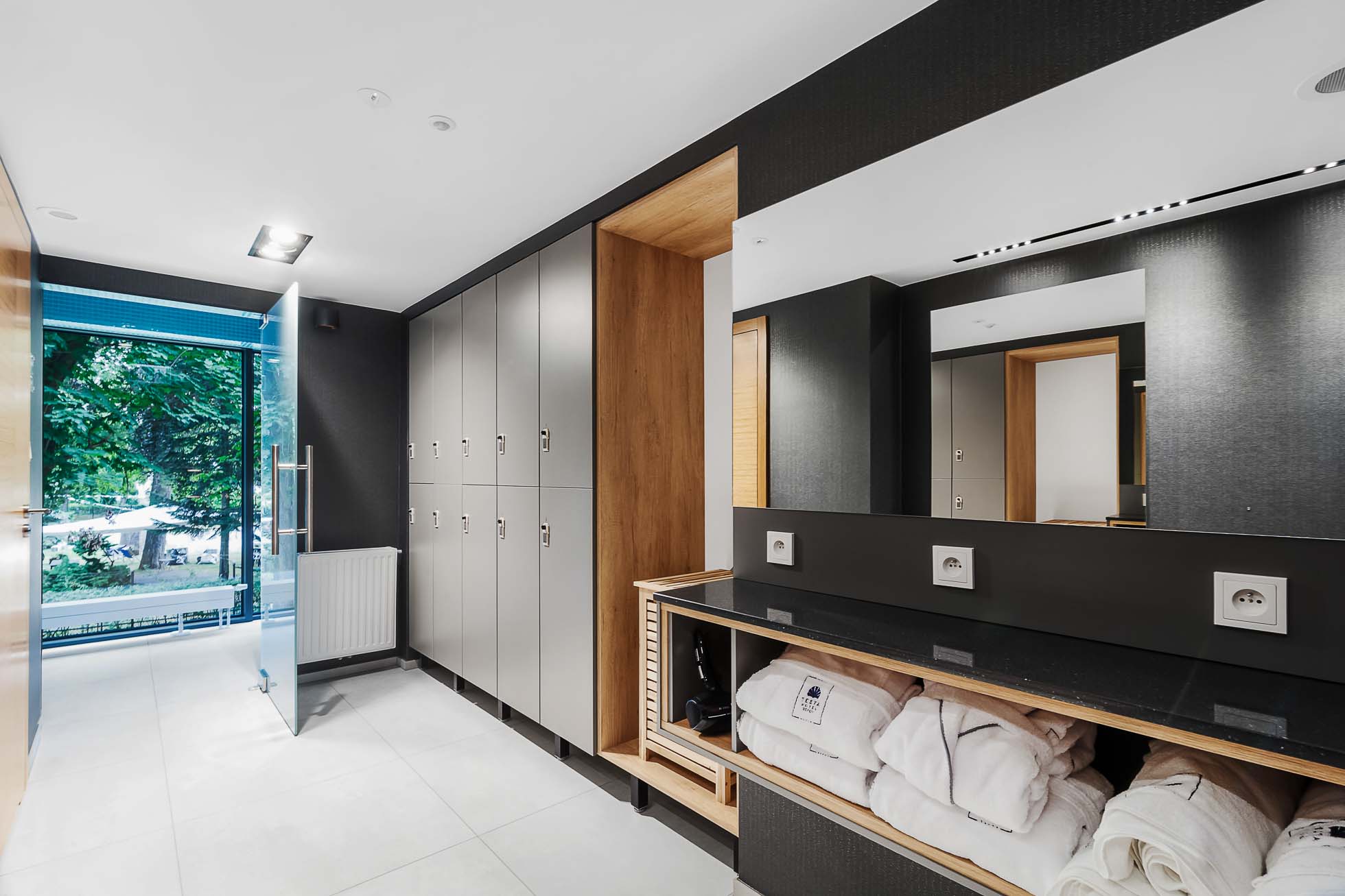 Nowoczesne spa w Sopocie z szafkami po lewej stronie i półkami na złożone ręczniki i szlafroki po prawej. Nad półkami znajduje się duże lustro. Otwarte drzwi prowadzą na teren zewnętrzny z zielenią widoczną przez szklane panele, prezentując elegancki, minimalistyczny wystrój idealny dla fotografia wnętrz Trójmiasto.