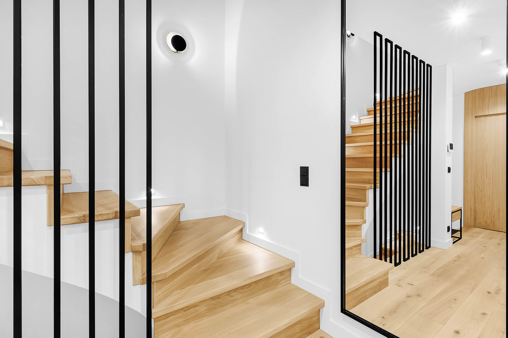 Nowoczesne, minimalistyczne wnętrze z drewnianymi schodami z eleganckimi, czarnymi metalowymi poręczami. Ściany są białe, a podłoga z jasnego drewna. Duże lustro na prawej ścianie podkreśla przestrzeń, odbijając schody i dodając elegancji pomieszczeniu. Idealne dla fotografia architektury Trójmiasto – zdjęcia wnętrz i budynków.