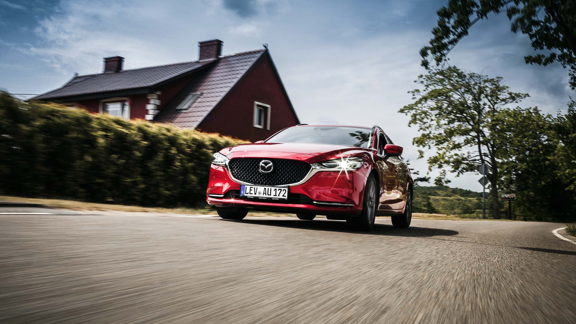 Czerwona Mazda sedan jedzie wiejską drogą z brązowym domem i zielonymi żywopłotami w tle, pod częściowo zachmurzonym niebem, uchwyconymi przez fotografia komercyjna i reklamowa Trójmiasto.