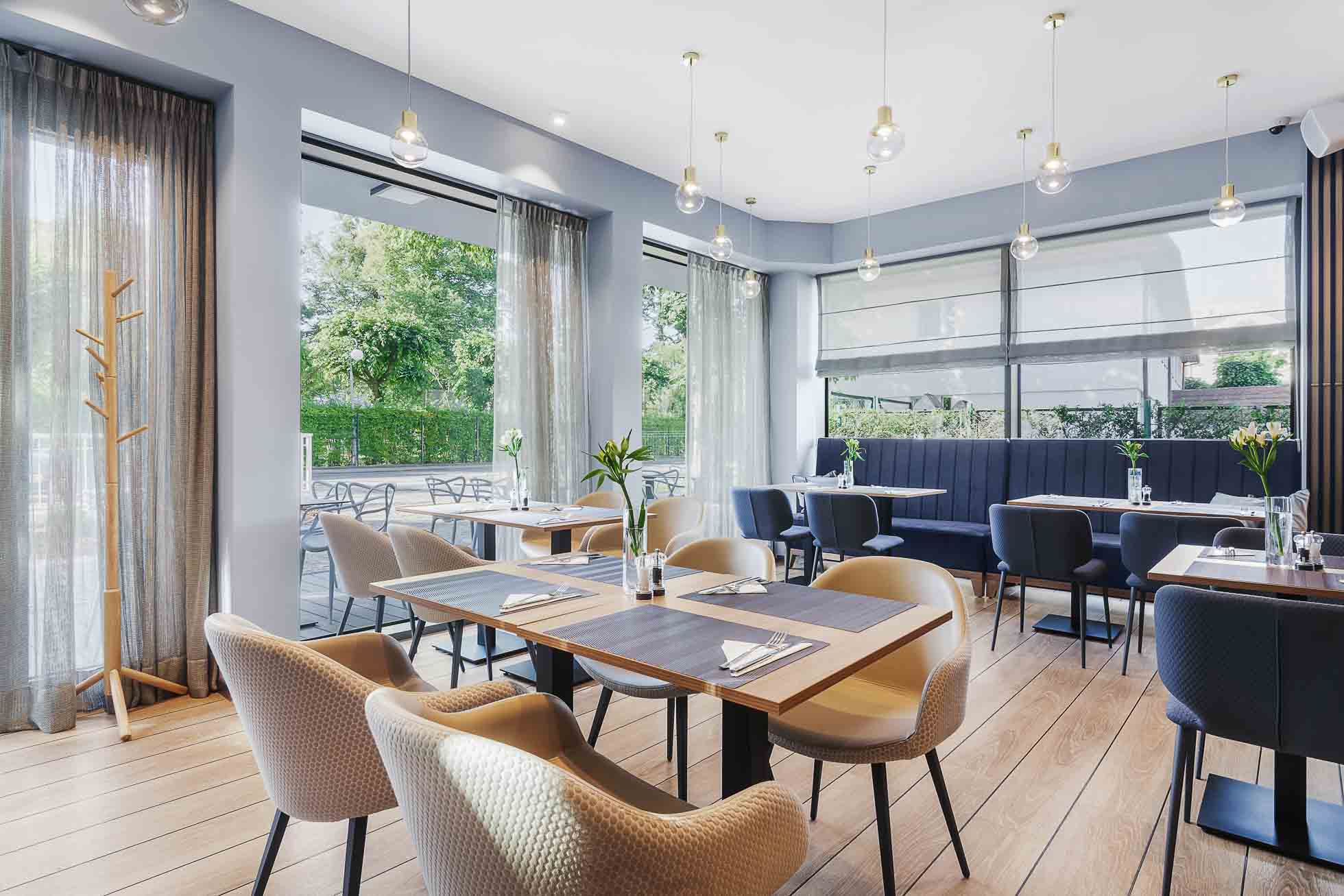 Jasne wnętrze restauracji z dużymi przeszkleniami, drewnianymi elementami i roślinnością